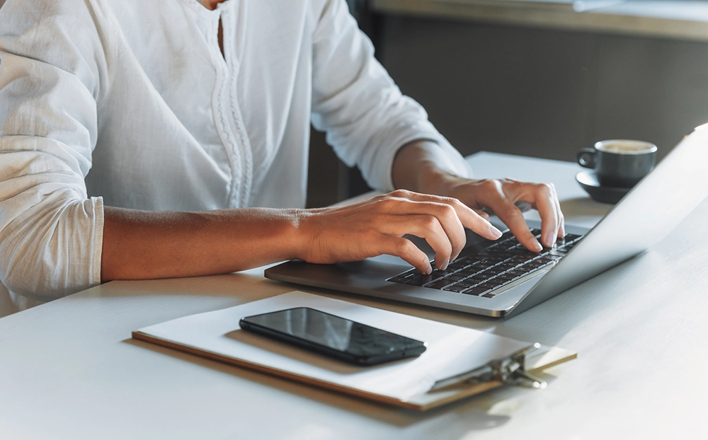 Une femme en tenue professionnelle utilise un ordinateur portable sur un bureau moderne. À côté, on aperçoit une calculatrice, un carnet, des stylos et une plante en pot, créant un environnement de travail structuré et organisé.