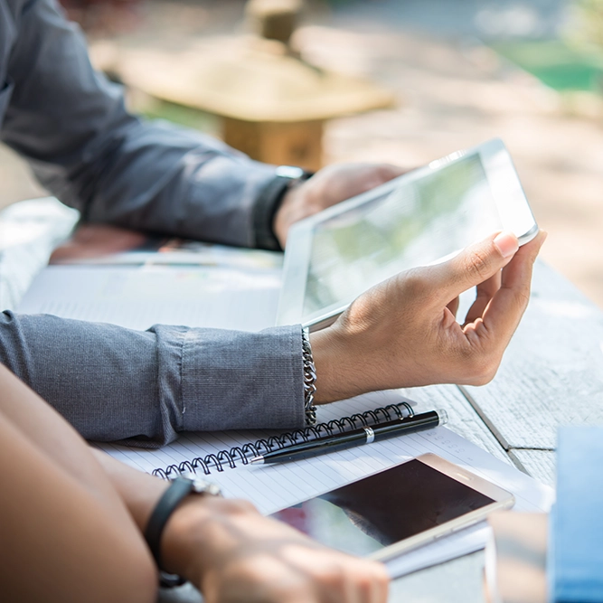 Deux personnes se retrouvent autour d'une table de travail en extérieur, utilisant un smartphone et une tablette tout en discutant. Des carnets et un stylo sont visibles en arrière-plan, mettant en valeur un environnement collaboratif.