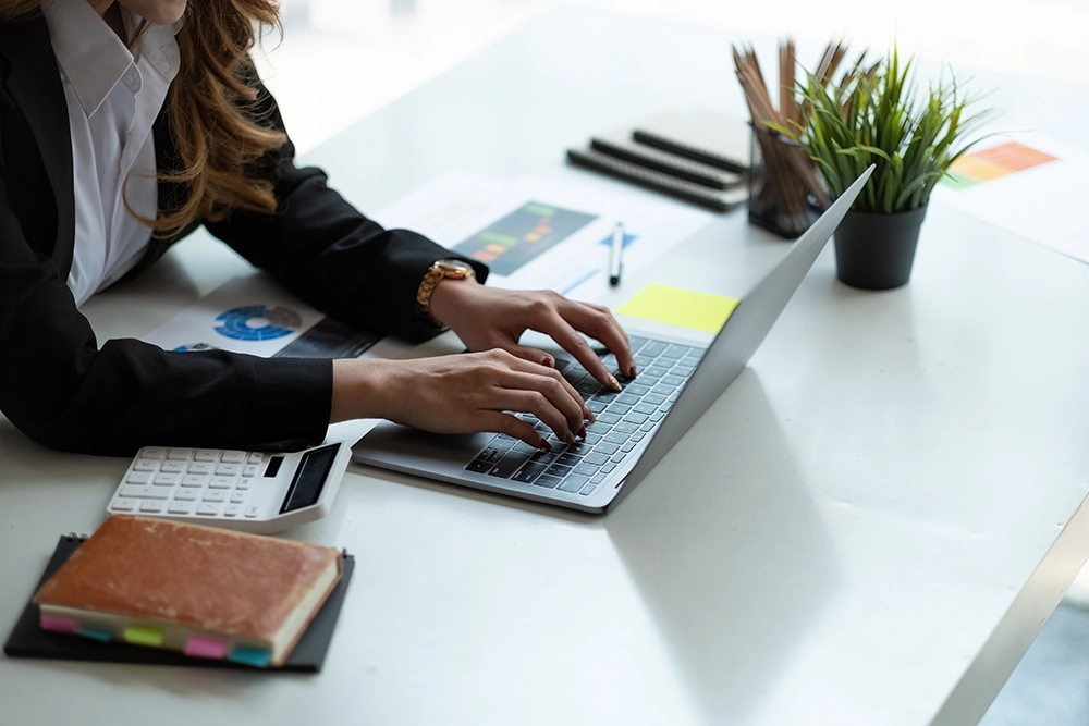 Une femme en tenue professionnelle utilise un ordinateur portable sur un bureau moderne. À côté, on aperçoit une calculatrice, un carnet, des stylos et une plante en pot, créant un environnement de travail structuré et organisé.